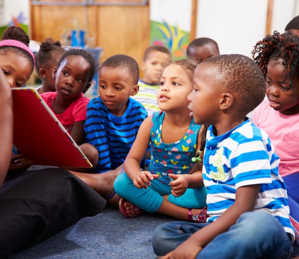 A group of preschoolers gathers around the carpet as their teacher reads them a book
