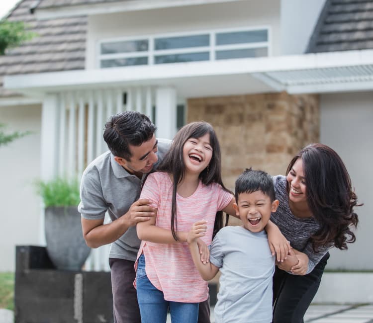 A family of four—two adults and two children—stands outside their house, smiling and laughing together, showing joy and affection.